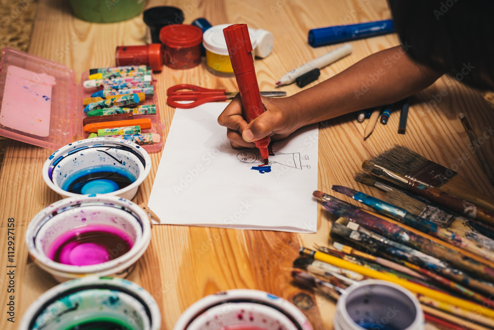 Child drawing at painting workshop. Painting tools on wooden board with sheet of white paper. Used colorful paintbrushes and crayons.
