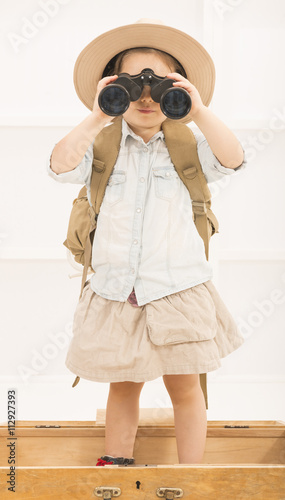 Adorable little explorer girl in a safari hat and explorer clothes with binoculars playing Safari sitting in a old wooden suitcase. Little child playing treasure hunt. Looking for the summer vacation