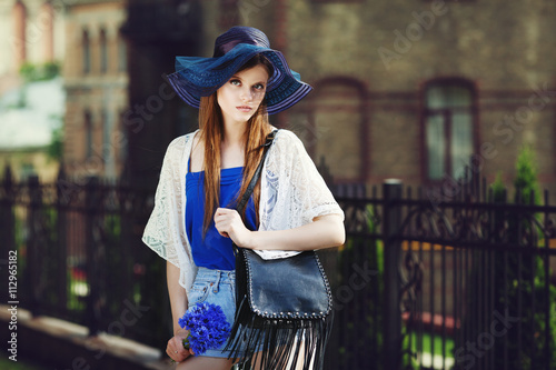 Outdoor portrait of young beautiful fashionable lady posing on street. Model wearing stylish summer clothes & wide-brimmed hat. Girl looking at camera. Sunny day. City lifestyle. Female fashion. Toned