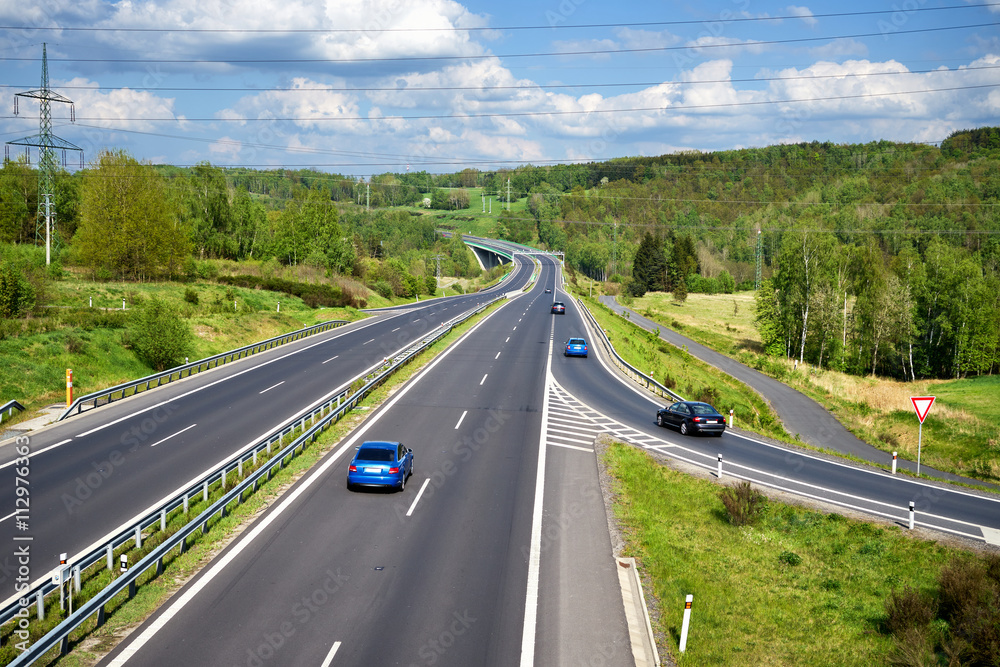 Row of blue cars and black cars entering the asphalt highway between lush forests in the countryside. In the distance the bridge and Electronic toll gate. Sunny day with white clouds in the blue sky.