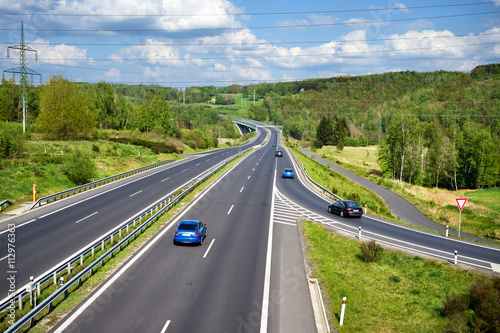 Obraz na płótnie Row of blue cars and black cars entering the asphalt highway between lush forests in the countryside. In the distance the bridge and Electronic toll gate. Sunny day with white clouds in the blue sky. w salonie