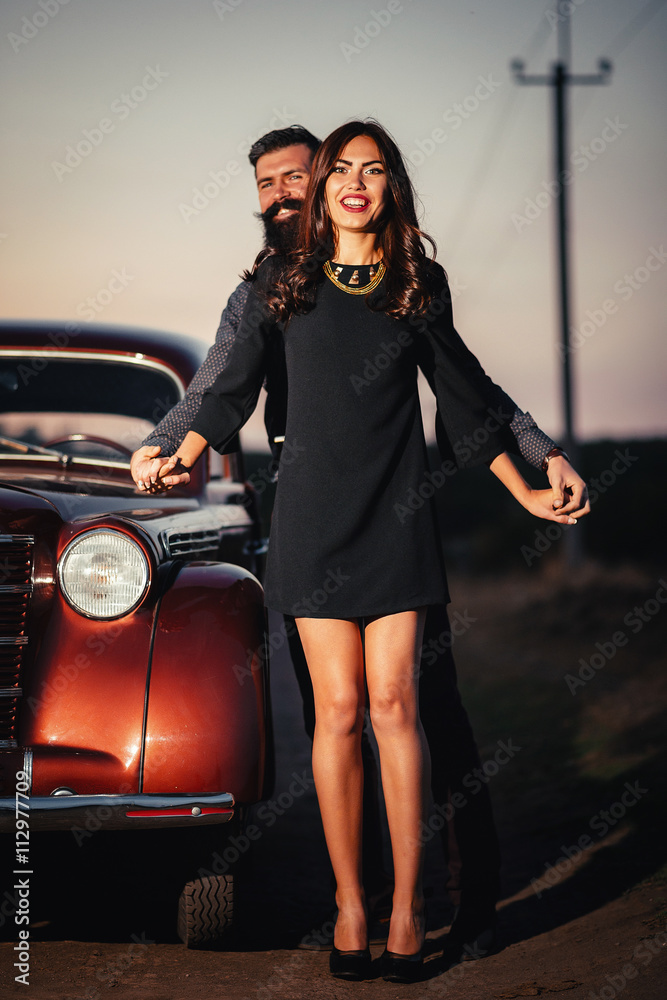 brutal bearded man with a mustache in a shirt, pants with suspenders with a girl with dark hair and big lips with bright red lipstick in a short dress and heels near retro car at sunset brutal bearded man with a mustache in a shirt, pants with suspenders with a girl with dark hair and big lips with bright red lipstick in a short dress and heels near retro car at sunset