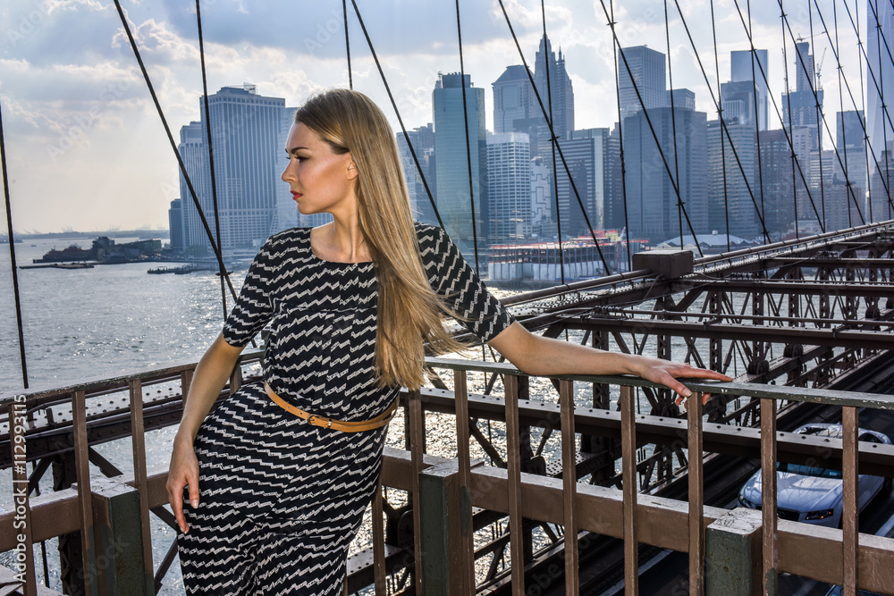 Attractive blonde young woman posing on the Brooklyn Bridge with city view on the background.
