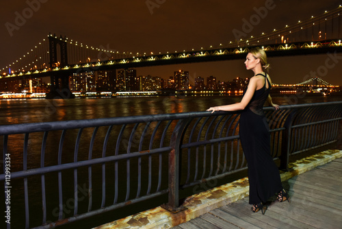 Fashion model posing sexy,wearing elegant evening dress at waterfront location with Manhattan bridge on the background. Night shot.