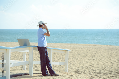 Busy male working on the laptop and talking on mobile. Man in hat speaking on smartphone over blue sky and ocean beach 