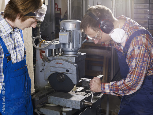 A man using an electric saw in a workshop while a young man looks on photo