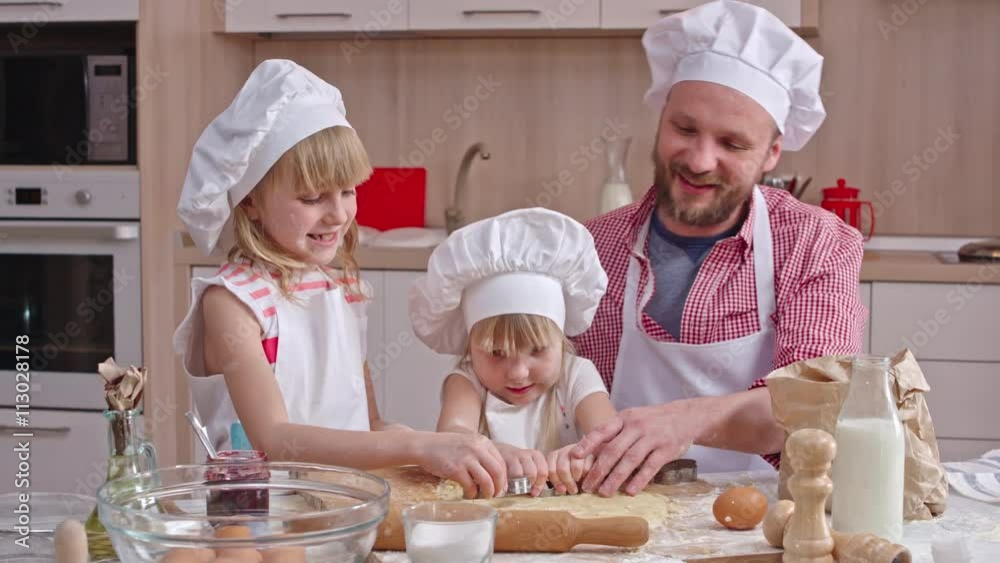 Lovely family spending their leisure time in the kitchen: dad and two little daughters enjoying cutting out cookies from dough together Lovely family spending their leisure time in the kitchen: dad and two little daughters enjoying cutting out cookies from dough together