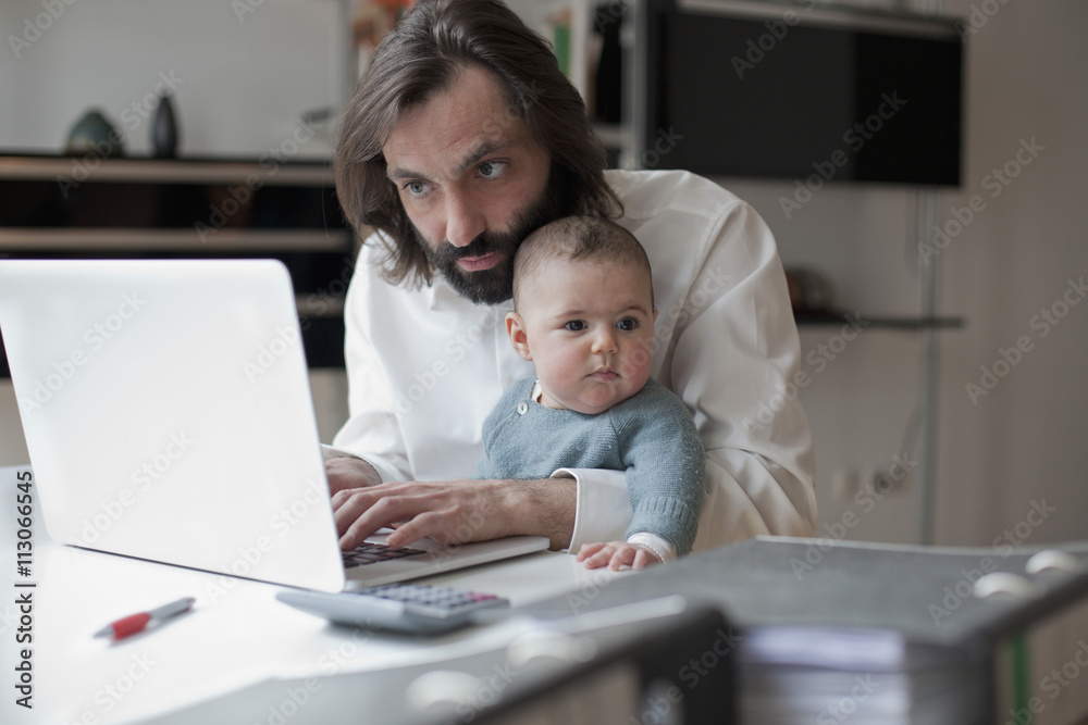 Mid adult man working on laptop while sitting with baby girl at home Mid adult man working on laptop while sitting with baby girl at home