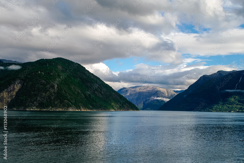 Cloudy sky over banks of fjord. Mountains with green forest, Eidfjord, Norway