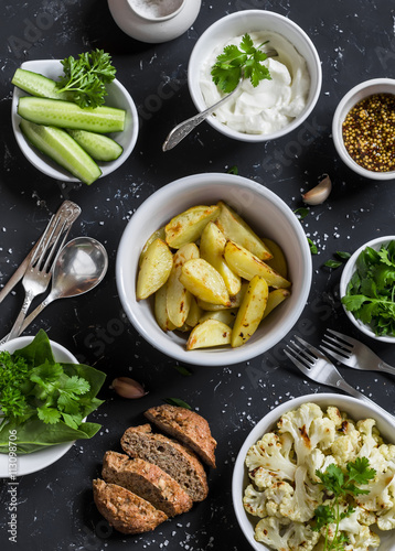 Baked cauliflower and potatoes, fresh cucumbers and herbs, sauces and spices on a dark stone background. Healthy vegetarian lunch or snack in a rustic style. Top view