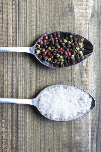 salt and pepper. Two metal spoons witlh salt crystals and color peppercorns on wooden table. photo