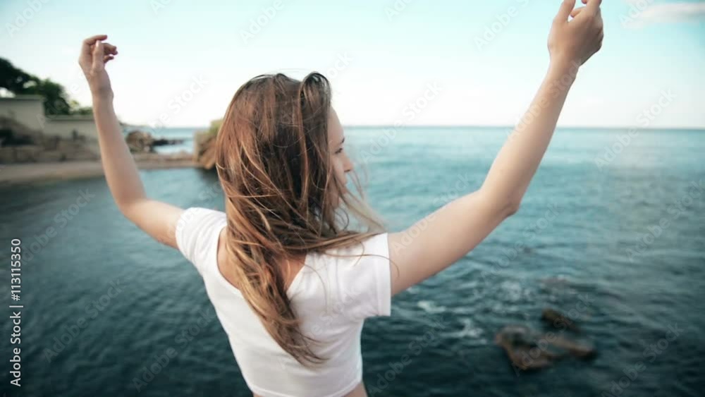 The young woman raised her hand up to the sun, against the sea, rocks and sky with clouds The young woman raised her hand up to the sun, against the sea, rocks and sky with clouds