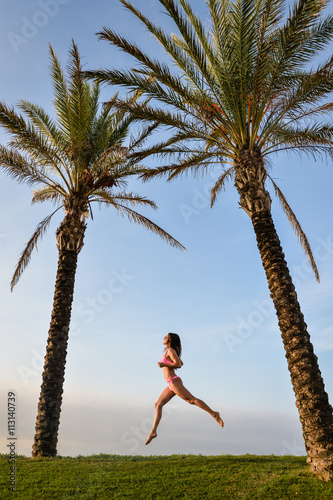 Picture of beautiful young excited lady in pink bikini dancing beside palm tree. Pretty girl with raised leg on summer blue sky background.