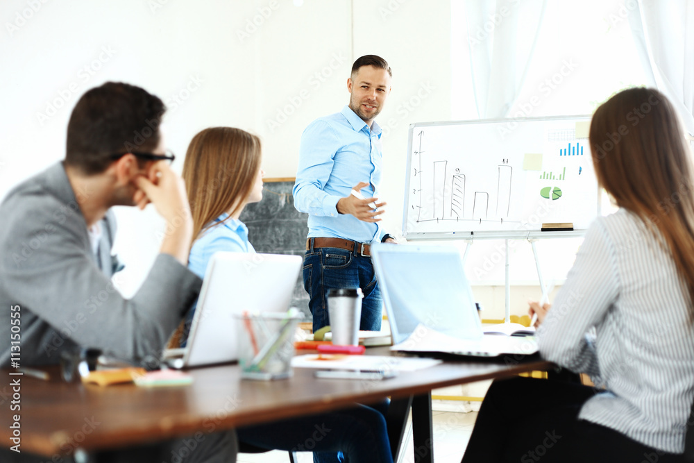 Handsome young man standing near whiteboard and pointing on the chart while his coworkers listening and sitting at the table Handsome young man standing near whiteboard and pointing on the chart while his coworkers listening and sitting at the table