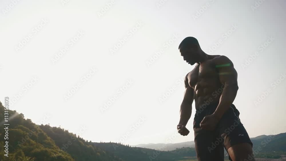 Silhouette of black african american handsome athlete man showing muscles on the rocky mountain background. Beauty and perfection of human's body