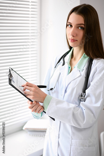 Female doctor brunette sitting at the table near the window in hospital and typing at laptop computer