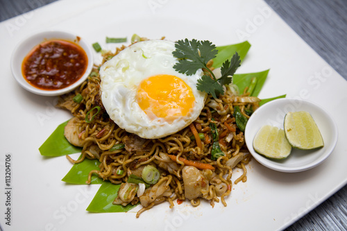 A delectable serving of Indonesian fried noodles (mie goreng) garnished with a squeeze of fresh lime, a spicy sambal condiment, and a perfectly cooked sunny-side-up egg. 