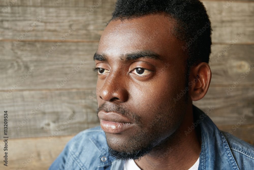 Obraz premium Highly-detailed profile of handsome young African man wearing denim jacket over white T-shirt looking with thoughtful and dreaming expression, making plans for the day, against wooden wall background
