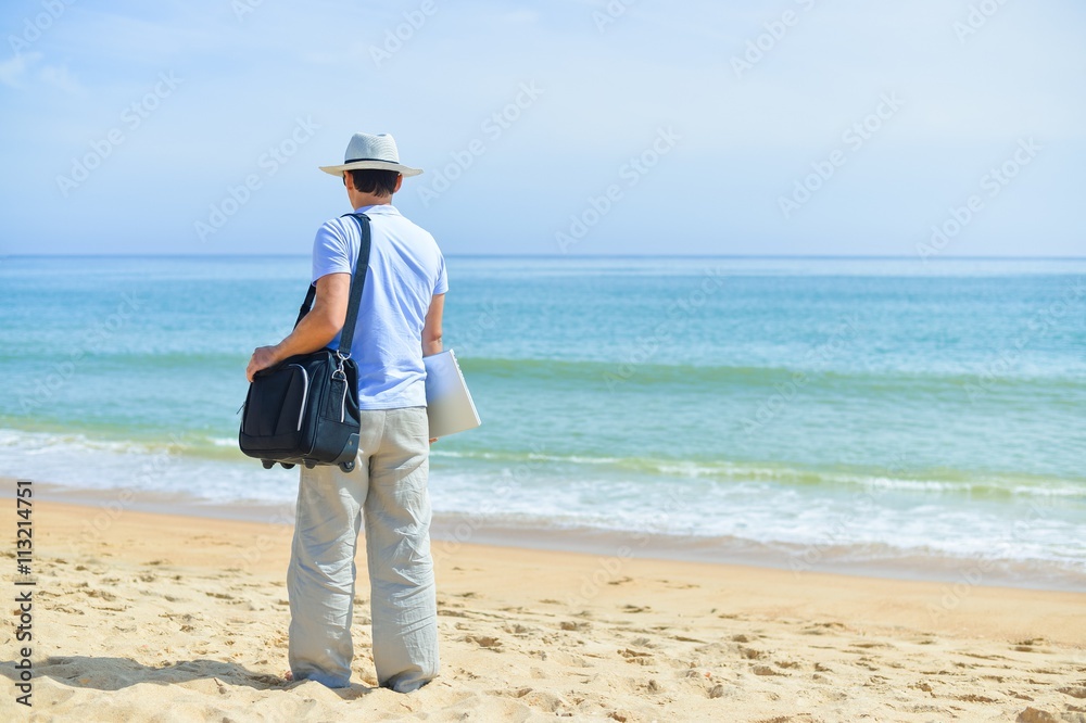 Back side view of man in hat holding travel bag on summer vacation over water and blue sky