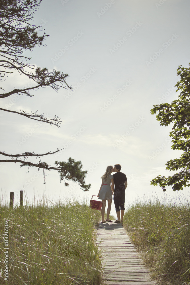 Rear view of couple kissing while standing on boardwalk amidst field against sky Rear view of couple kissing while standing on boardwalk amidst field against sky