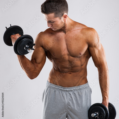 Muscular bodybuilder guy doing exercises with dumbbells over grey background with copyspace