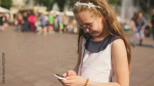 Blond kid girl playing with smartphone sitting on park lawn in city skyline background