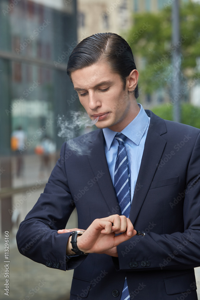 Junger Mann im Businessstil, Anzug, Hemd und Krawatte raucht E-Zigarette in einer modernen Stadtkulisse und schaut dabei auf seine Armbanduhr Junger Mann im Businessstil, Anzug, Hemd und Krawatte raucht E-Zigarette in einer modernen Stadtkulisse und schaut dabei auf seine Armbanduhr