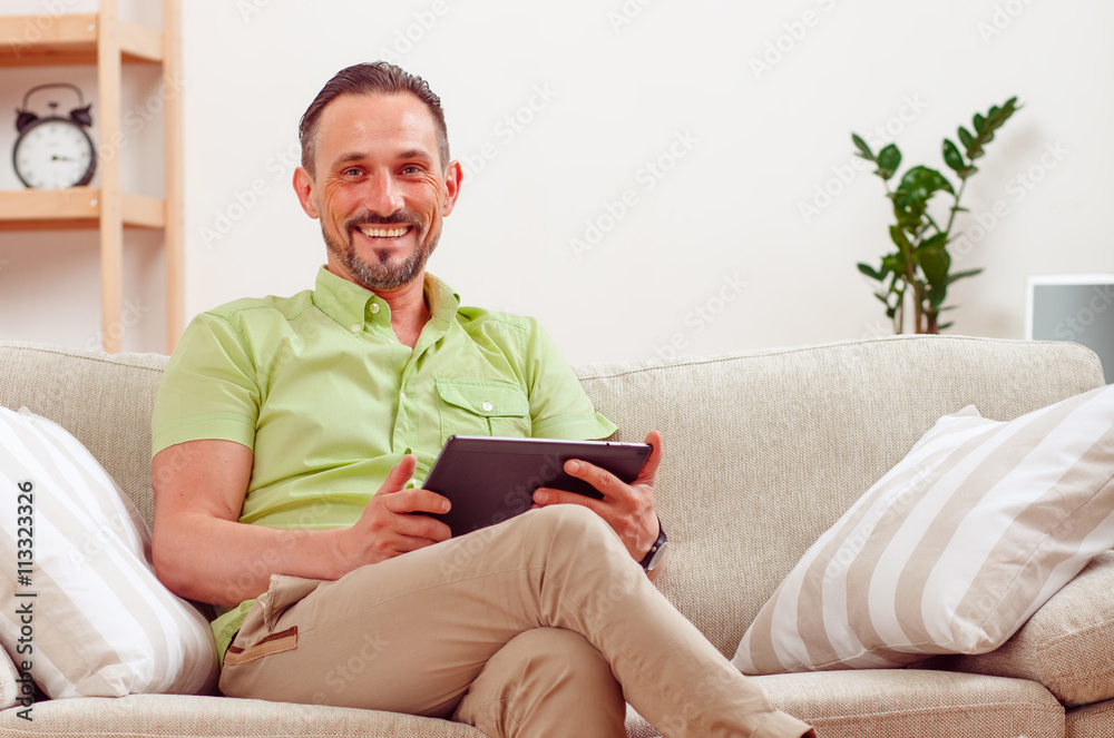 Portrait of handsome businessman using tablet PC at home. Happy man sitting on safa or couch and working from home.