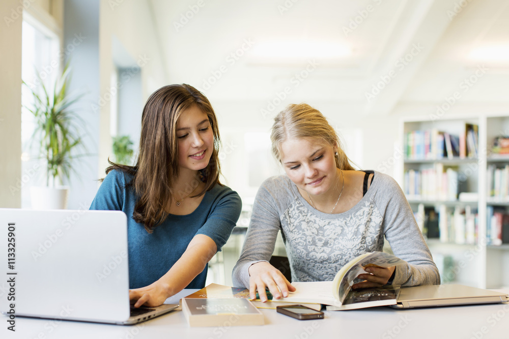 Teenage girls reading book while using laptop at table in school library Teenage girls reading book while using laptop at table in school library