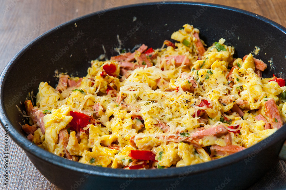scrambled eggs with ham, vegetables and cheese in a frying pan on wooden table scrambled eggs with ham, vegetables and cheese in a frying pan on wooden table