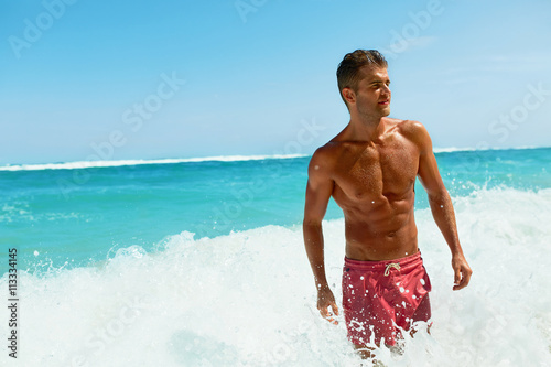 Sexy Man On Beach In Summer. Handsome Male With Fit Body, Healthy Skin Sun Tan Coming Out Of Sea At Luxury Relax Spa Resort. Beautiful Happy Guy Relaxing, Enjoying Holidays Travel Vacation. Summertime