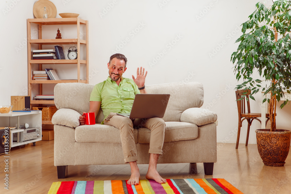 Portrait of handsome businessman waving while sitting on sofa or couch, drinking coffee or tea and working on laptop computer.