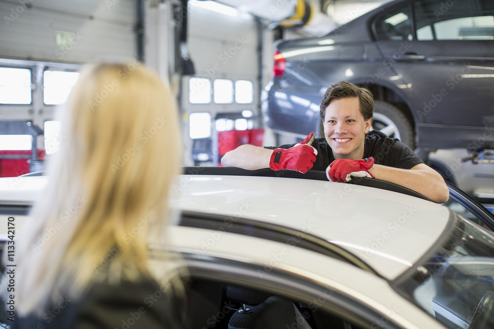 Confident young mechanic showing thumbs up to female customer over car Confident young mechanic showing thumbs up to female customer over car