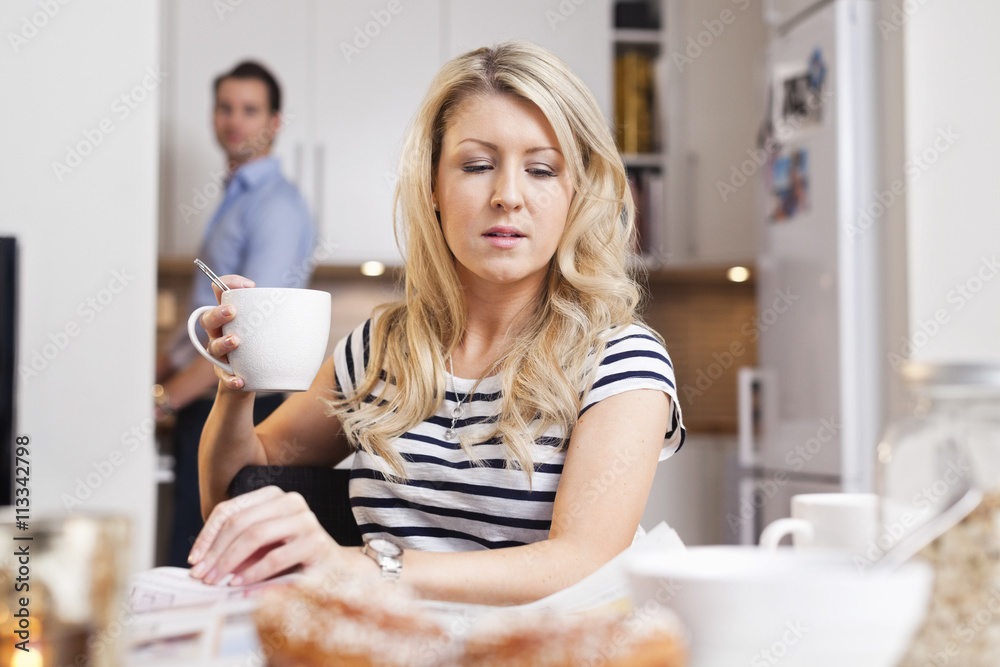 Young woman holding coffee cup reading newspaper with man standing in background Young woman holding coffee cup reading newspaper with man standing in background