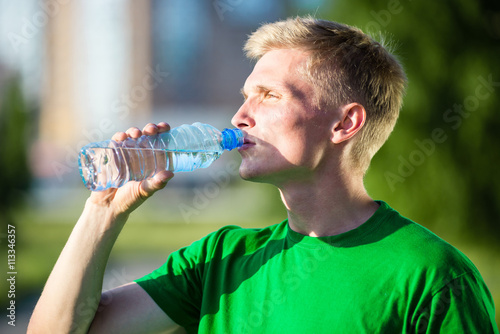 Tired man drinking water from a plastic bottle after fitness time and exercising in city street park at beautiful summer day. Sporty model caucasian ethnicity training outdoor. 