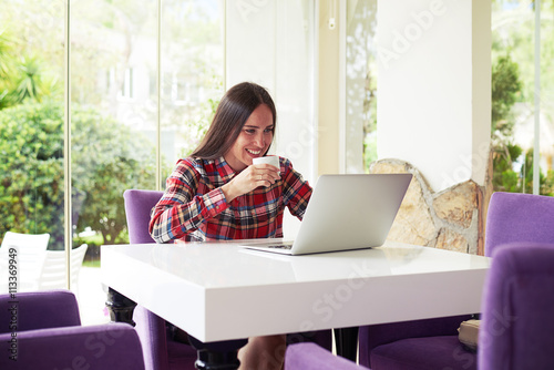 Young dark-haired woman enjoys coffee while working with laptop