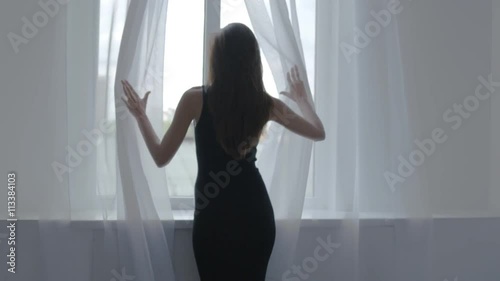 Woman in black dress stands near window and opens white curtains, back to camera
