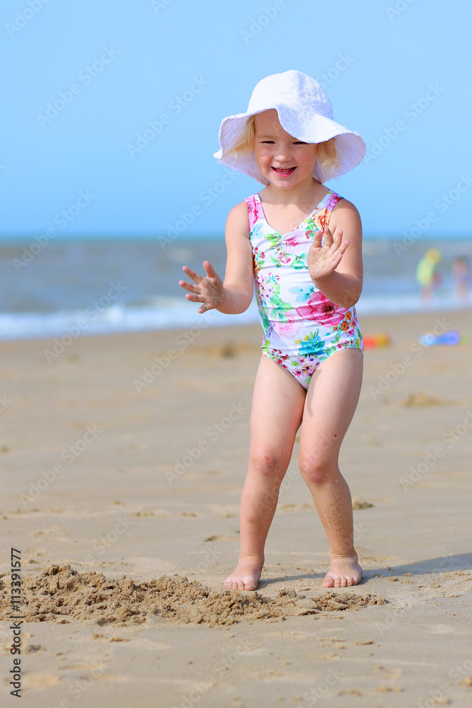 Cute active child wearing white hat playing on sandy beach. Happy little girl enjoying summer holidays on a sunny day. Family with young kids on vacation at the North Sea coast.