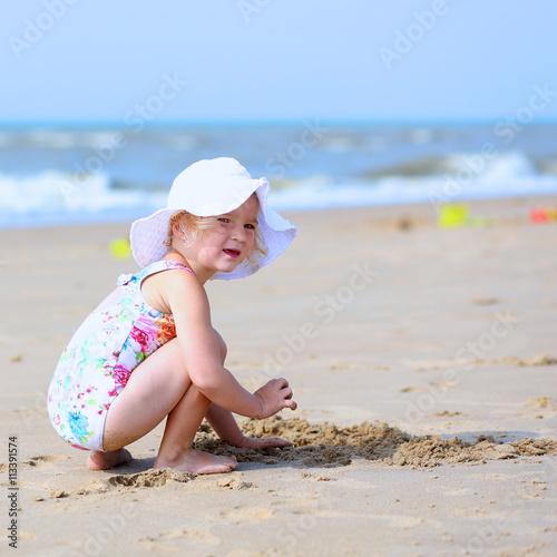 Cute active child wearing white hat playing on sandy beach. Happy little girl enjoying summer holidays on a sunny day. Family with young kids on vacation at the North Sea coast.