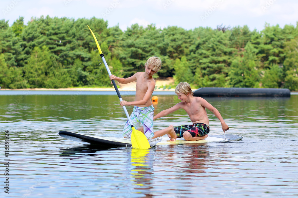 Adventurous boys learning to paddle on stand up board. Happy children, teenage schoolboys, having fun enjoying adventure experience on the river or lake on a sunny day during summer holidays