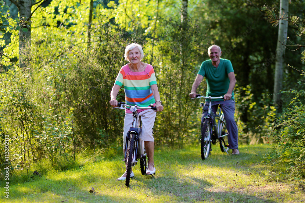 Fototapeta premium Happy healthy couple biking together in the forest. Seniors enjoying sunny day outdoors. Active retirement concept.