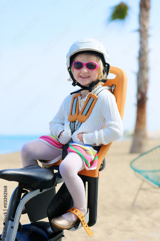 Naklejka premium Happy little girl enjoying ride on the bike. Toddler sitting in child bicycle seat. Family having fun outdoors. Beach promenade and sea at the background.