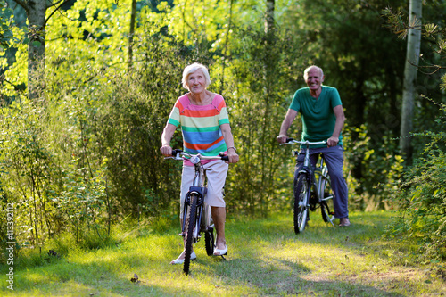Happy healthy couple biking together in the forest. Seniors enjoying sunny day outdoors. Active retirement concept. © cromary