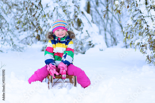 Portrait of beautiful toddler girl playing outdoors with snow. Happy little child wearing colorful knitted hat and blue coat enjoying winter day in the park or forest.