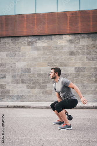 Fitness male crouching wile working out with kettlebell. Sporty man exercising outdoor on the street.