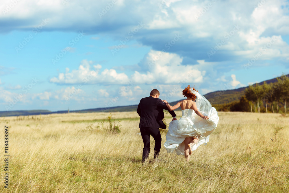 Bride jumps up walking behind a groom acroos a golden field Bride jumps up walking behind a groom acroos a golden field