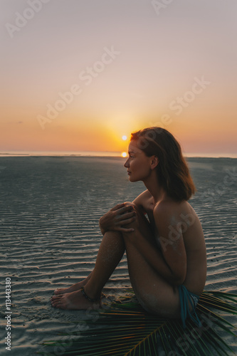 Young pretty girl sitting on the sand during sunset with palm branch