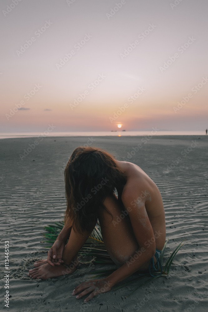 Young pretty girl sitting on the sand during sunset with palm branch Young pretty girl sitting on the sand during sunset with palm branch