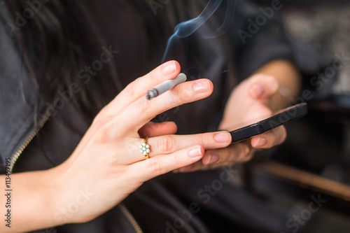 Young girl sitting in cafe  smoking cigarette and typing on smart phone  hands detail