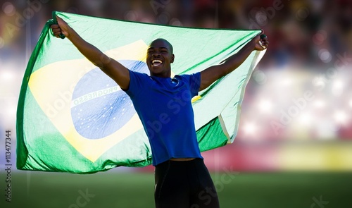 Front view of Brazilian sportsman is smiling and raising a flag  against sports arena